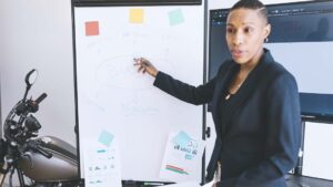 An businessperson of African ancestry points at a whiteboard while giving a presentation